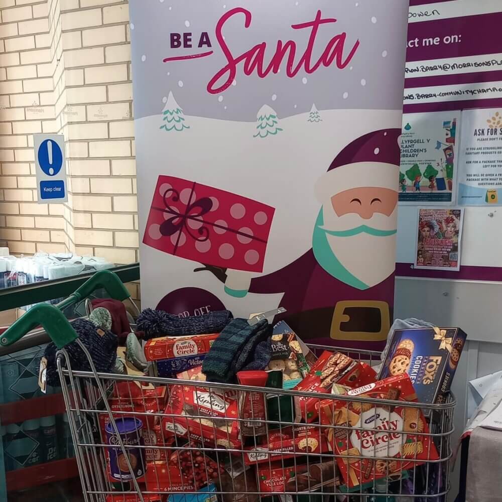 Shopping cart filled with holiday snacks in front of "Be a Santa" banner with Santa holding a gift. - Home Instead