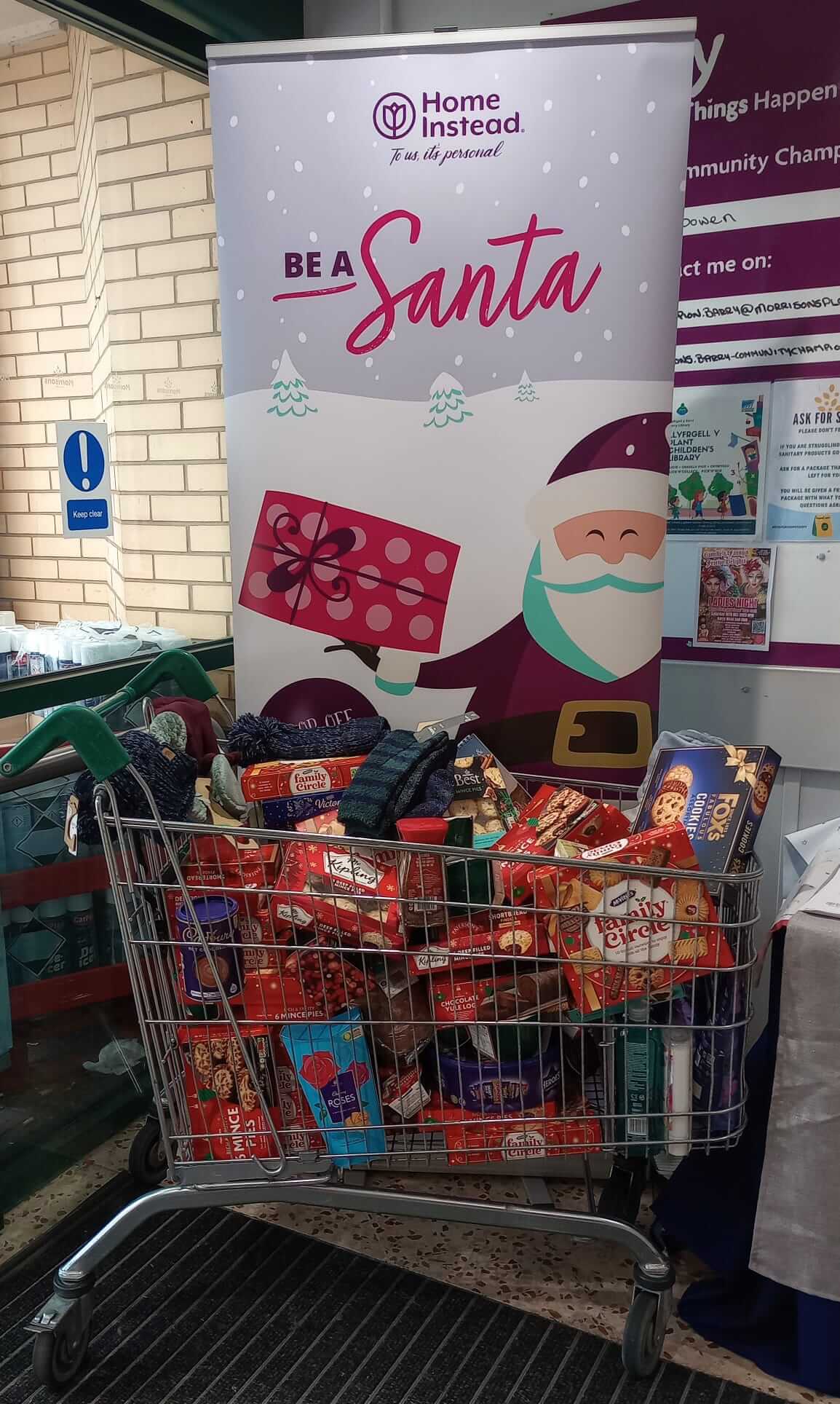 A shopping cart filled with various food items in front of a "Be a Santa" banner. - Home Instead