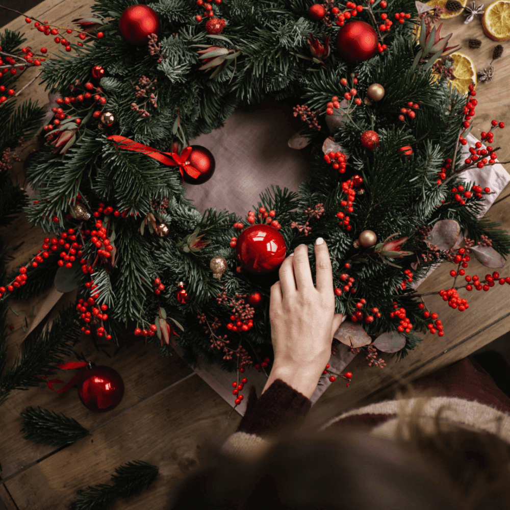 A hand placing a red ornament on a festive Christmas wreath decorated with red berries, pine cones, and green foliage. - Home Instead