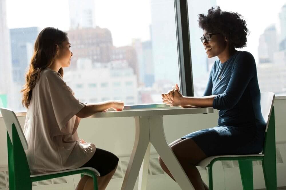 Two women sitting at a small table by a window, engaged in a conversation in a bright, modern setting. - Home Instead