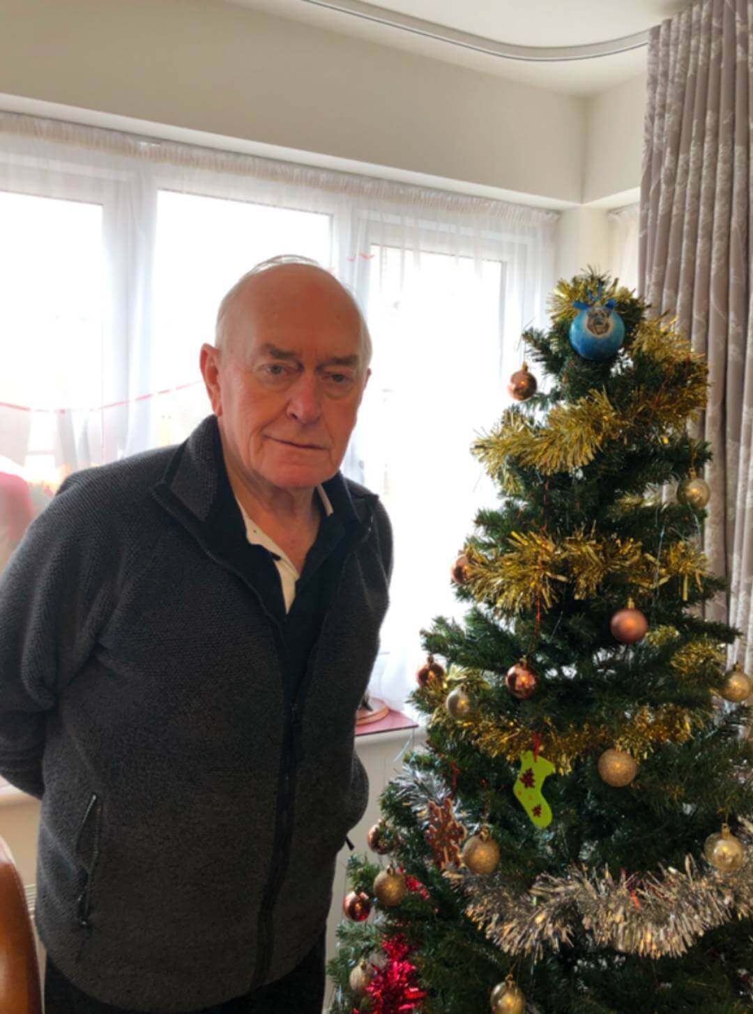 Elderly man stands next to a decorated Christmas tree in a well-lit room with large windows. - Home Instead