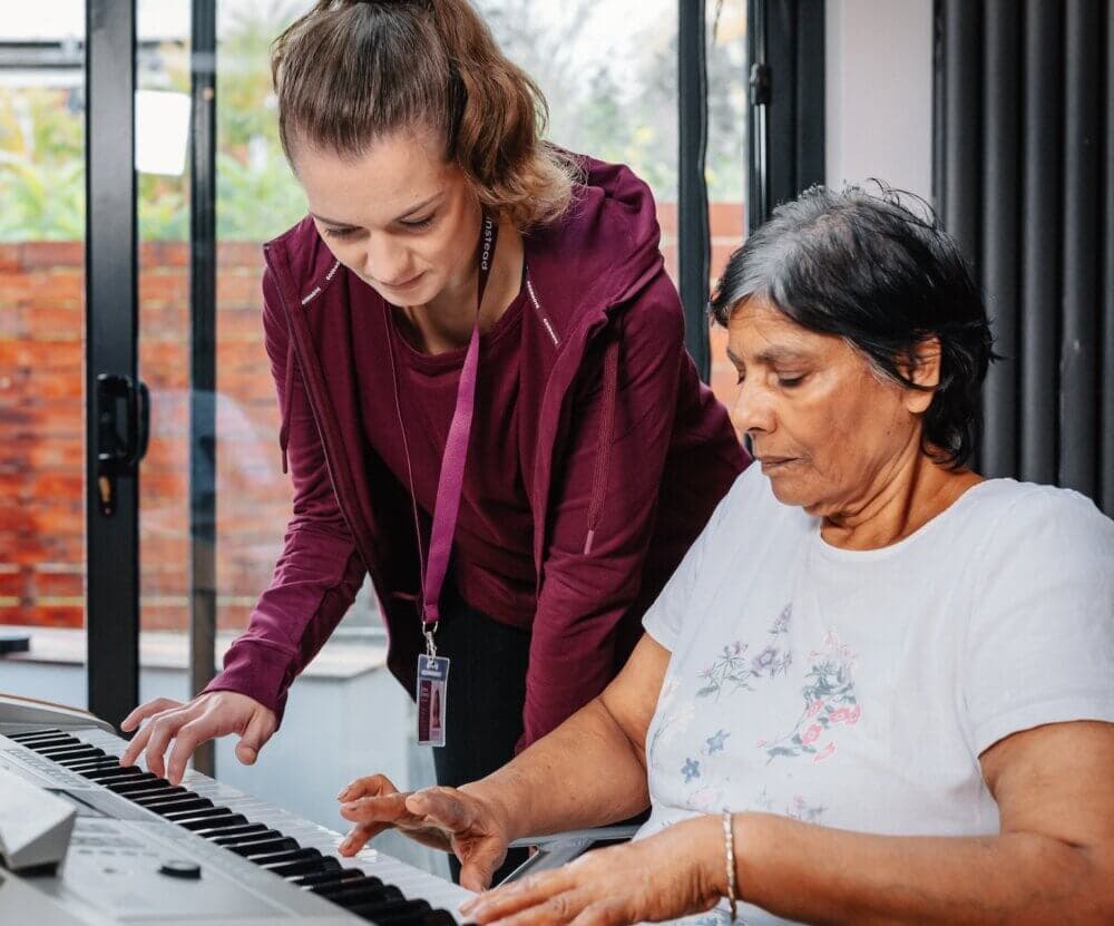 A woman guides an older woman playing a keyboard in a bright room with large windows. - Home Instead