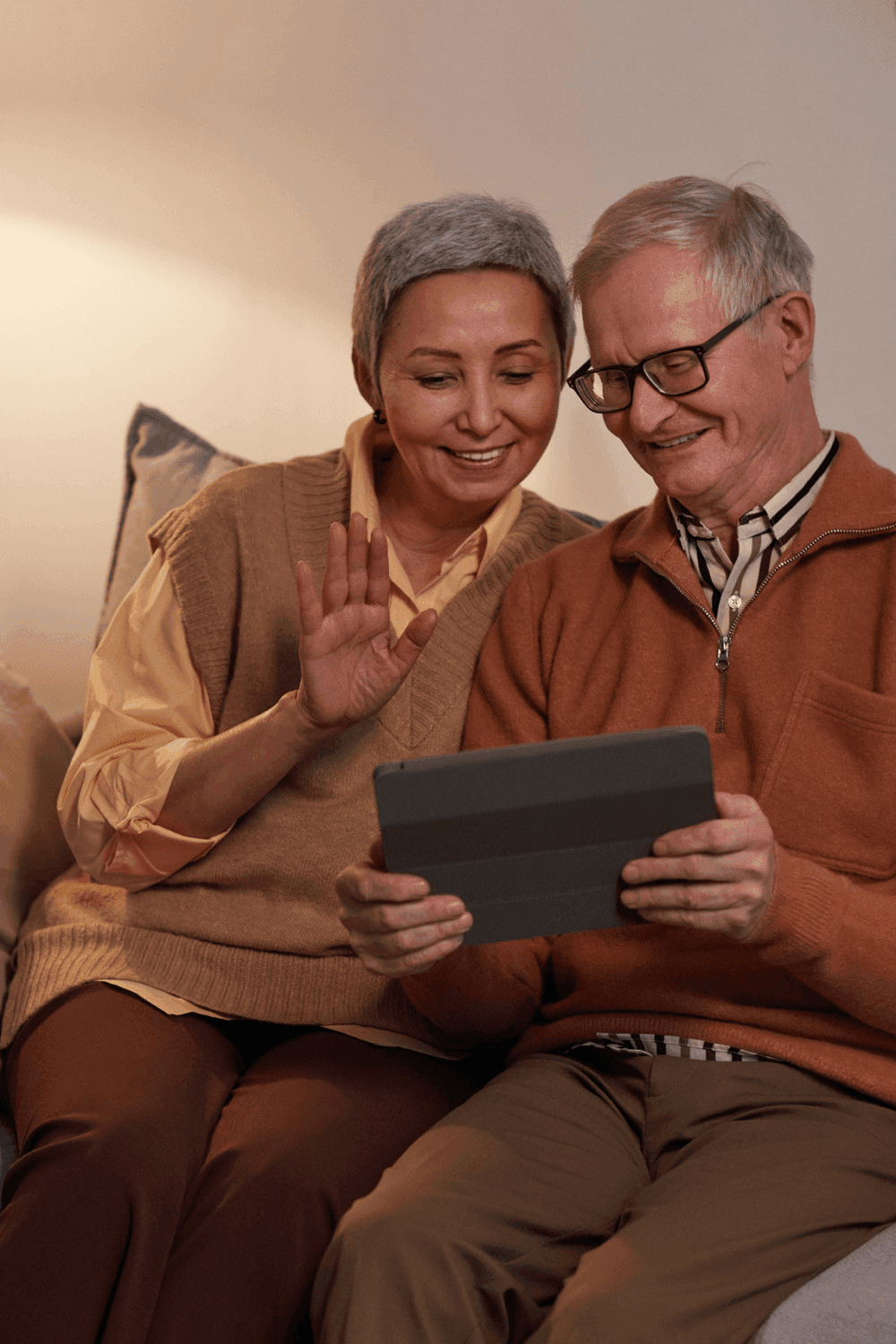 An elderly couple smiling and waving at a tablet screen while sitting together on a couch. - Home Instead
