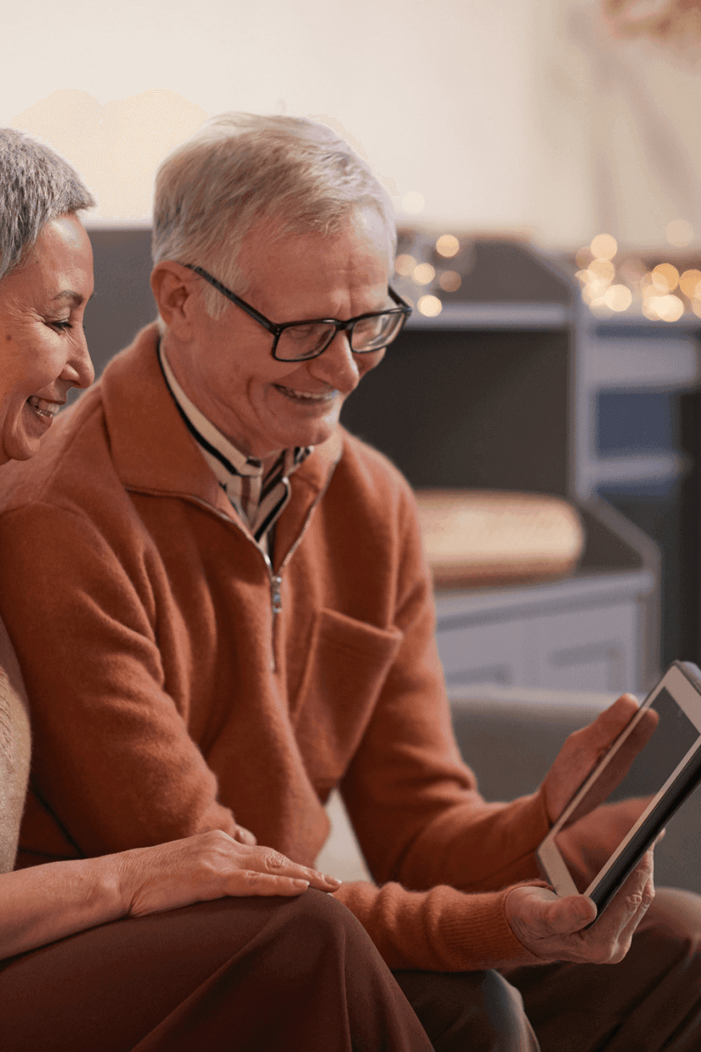 Two elderly individuals sitting close together, smiling and looking at a tablet in a cozy, warmly lit room. - Home Instead