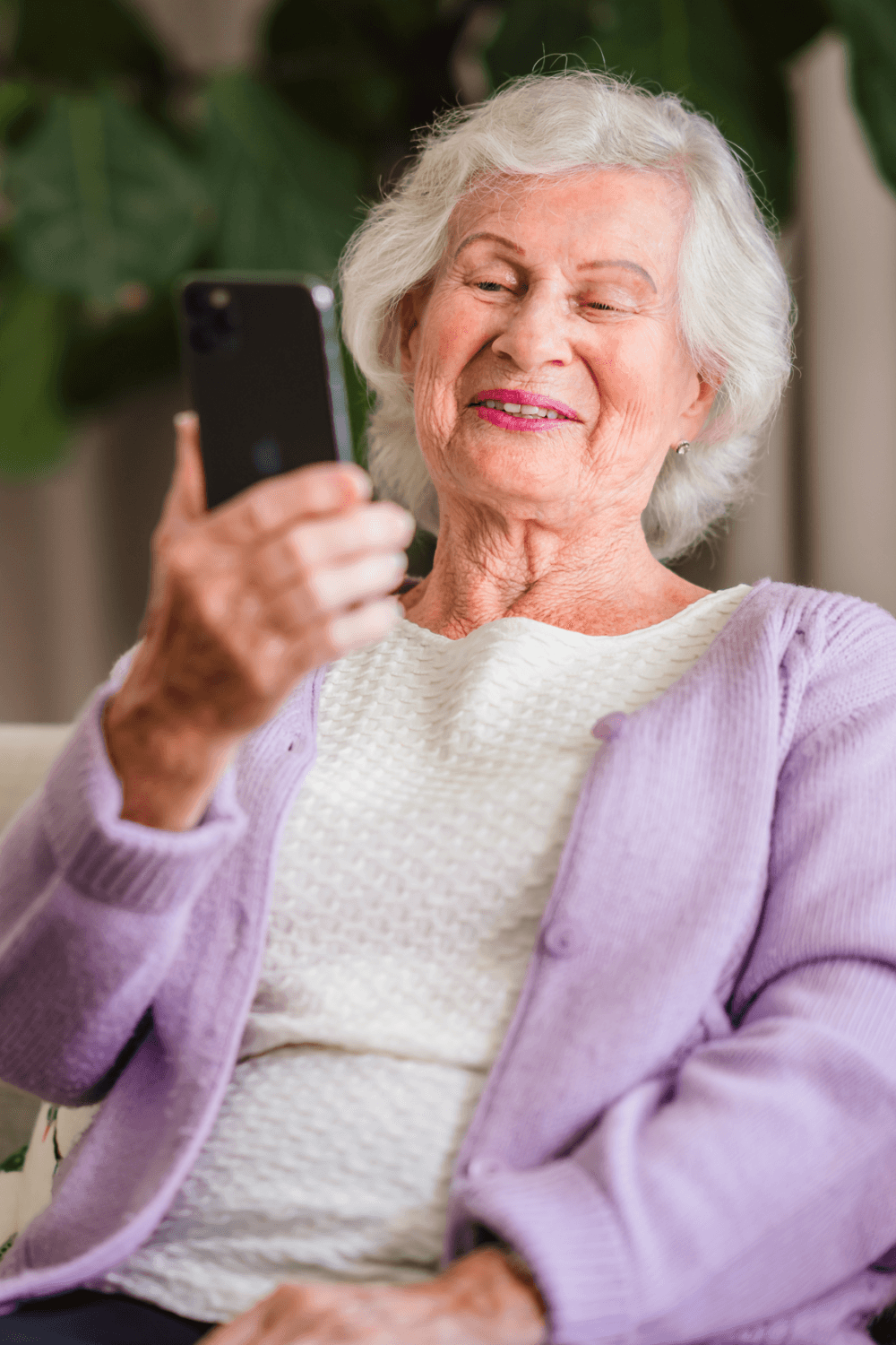 Elderly woman with white hair and a purple cardigan smiling while looking at a smartphone. - Home Instead