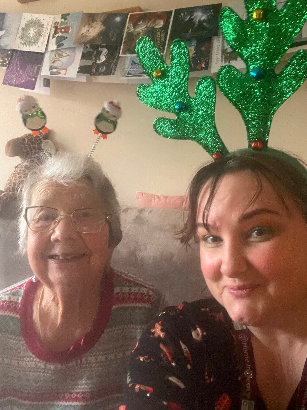 Two people sitting together, one older woman in festive sweater and a younger woman wearing sparkly green antler headband. - Home Instead