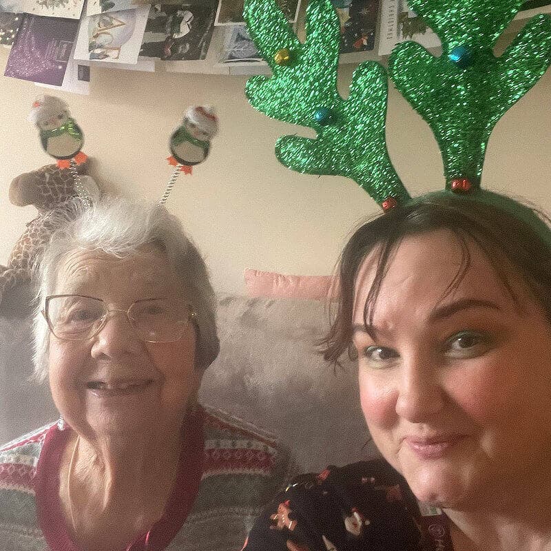 Two people wearing festive headbands with reindeer antlers and snowmen smile for a selfie in a room with holiday decor. - Home Instead