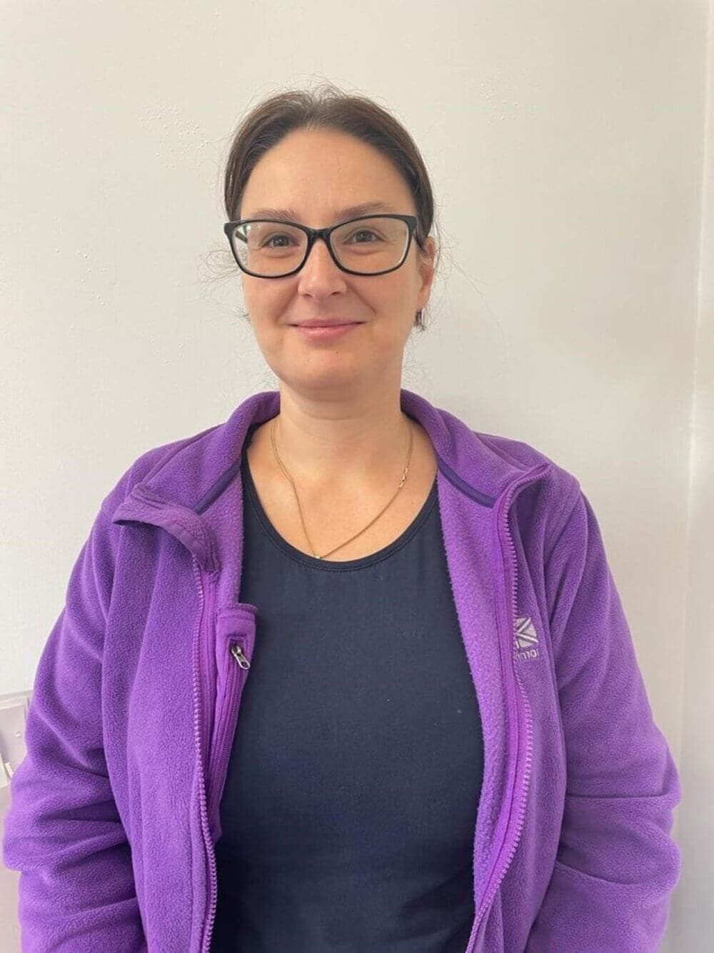 A woman with glasses and a purple jacket stands against a plain white background, smiling slightly. - Home Instead