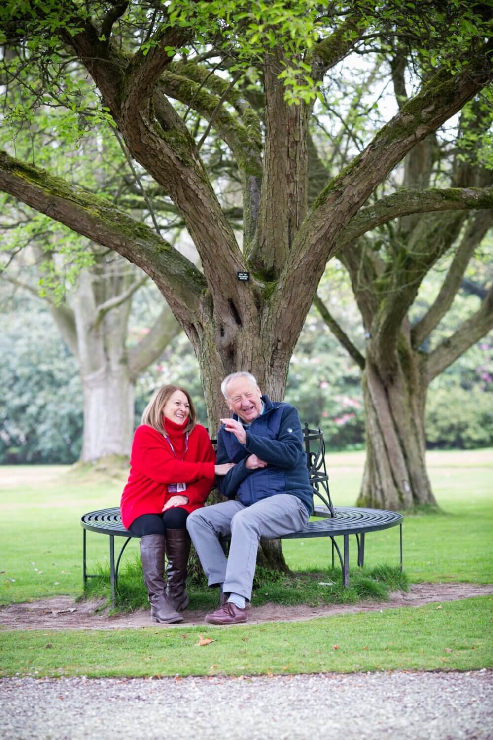 An elderly man and woman seated on a curved bench under a large tree, smiling and laughing together in a park. - Home Instead