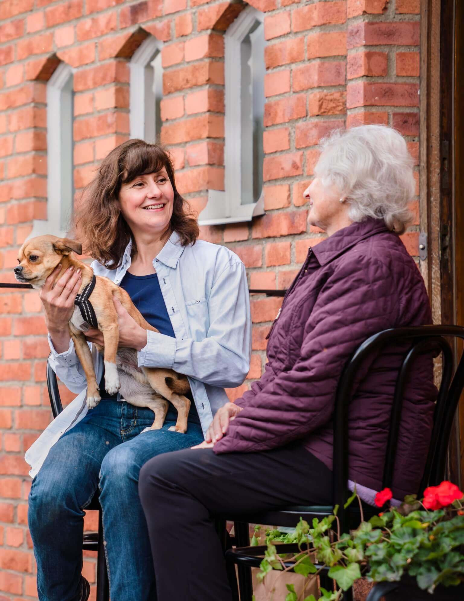 Two women seated outside by a brick wall, one holding a small dog, they are smiling and conversing. - Home Instead