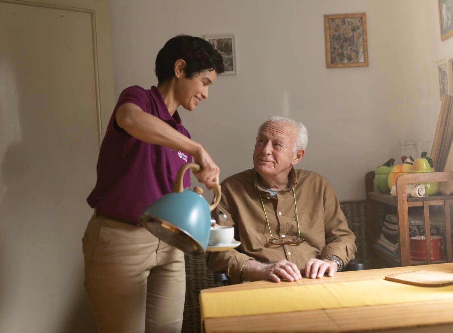 A caregiver in a purple shirt serves tea to an elderly man sitting at a table in a cozy room. - Home Instead