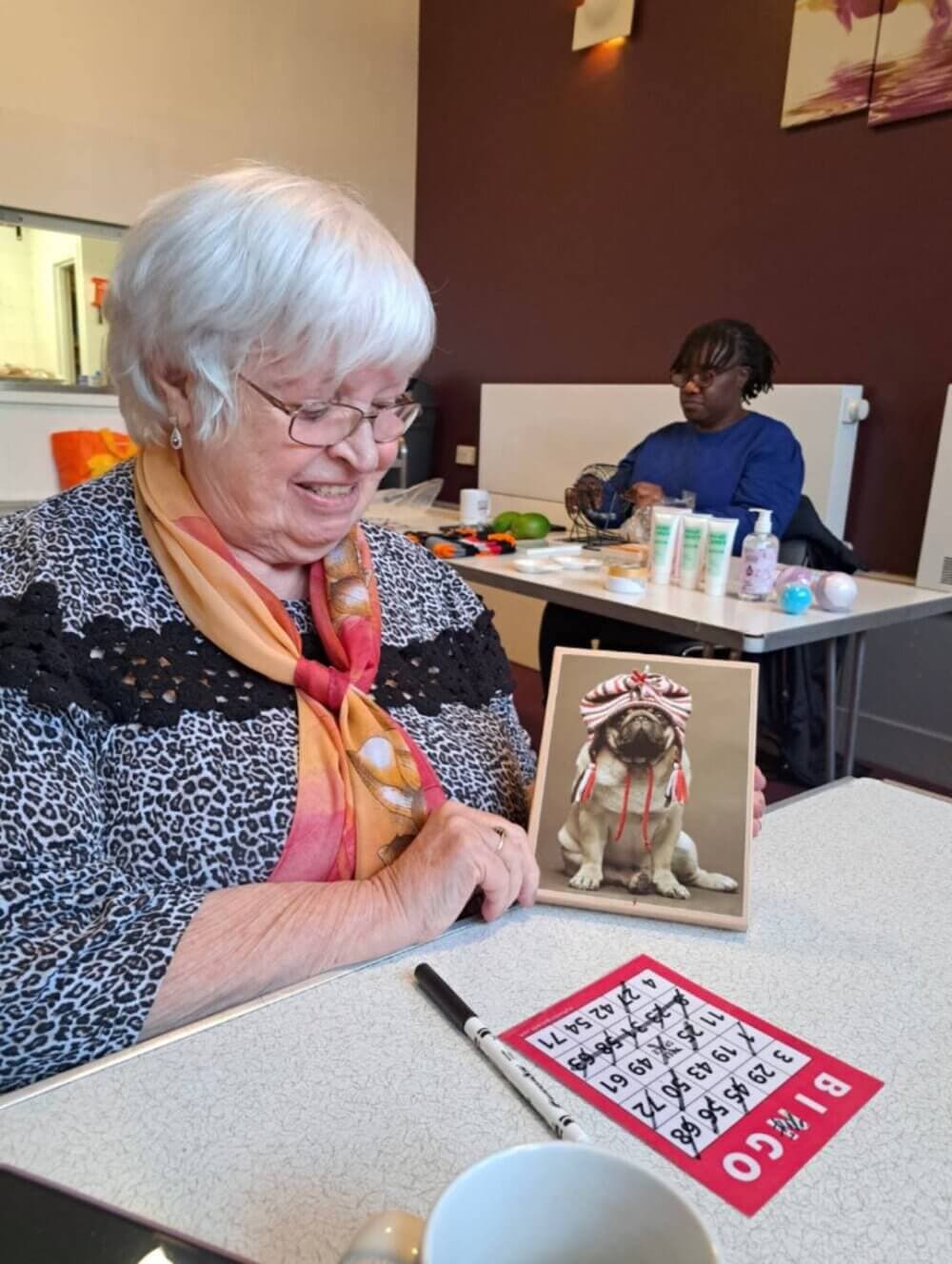 Elderly woman smiling at a greeting card with a dog, while sitting at a table with a bingo card in front of her. - Home Instead