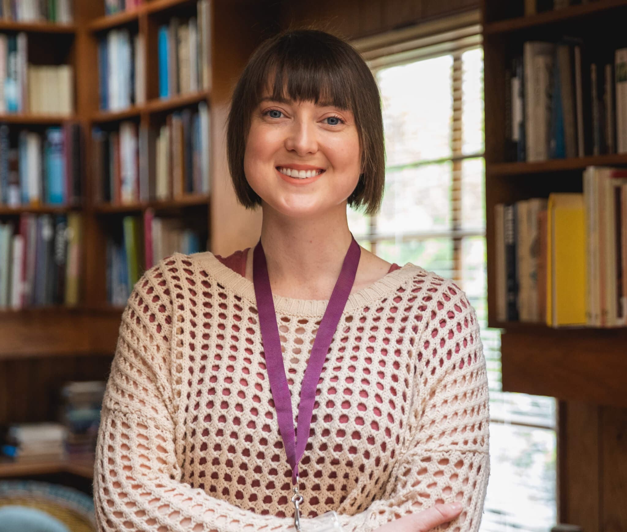Smiling person with short hair and a lanyard, in a cozy room with wooden shelves filled with books. - Home Instead