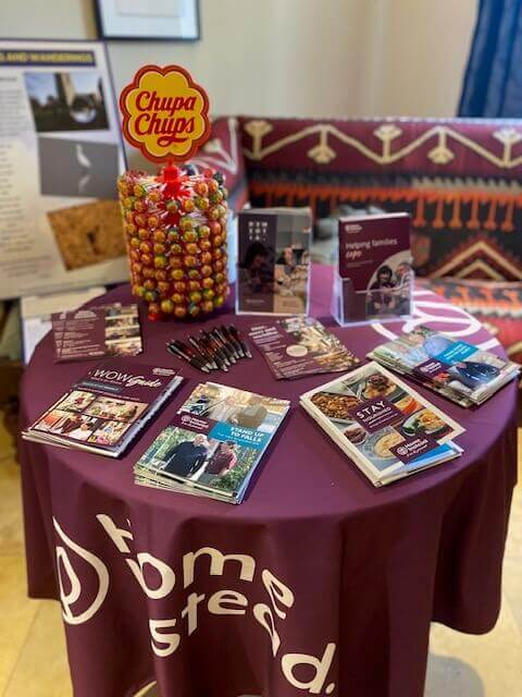 A round table with Chupa Chups lollipops, brochures, and pens, covered with a purple "Home Instead" tablecloth. - Home Instead