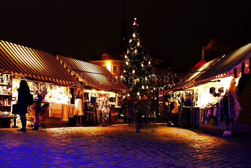 A festive Christmas market at night with decorated stalls, a large Christmas tree, and a busy cobblestone street. - Home Instead