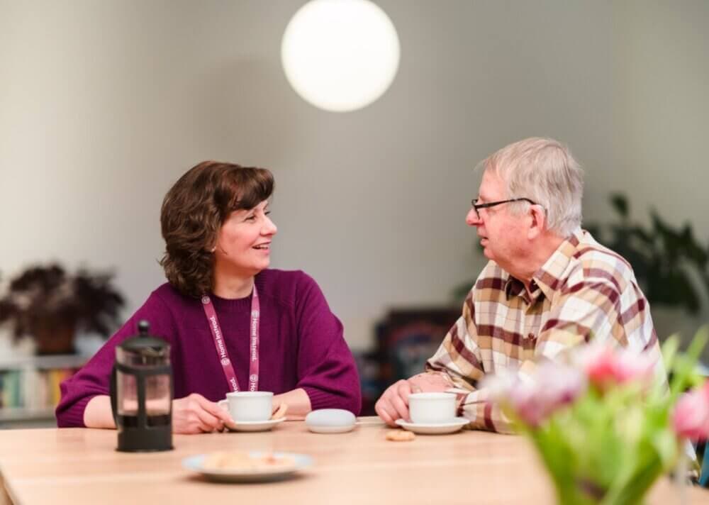 Two people sitting at a table in conversation, each holding a cup of tea or coffee, with a bright indoor setting. - Home Instead