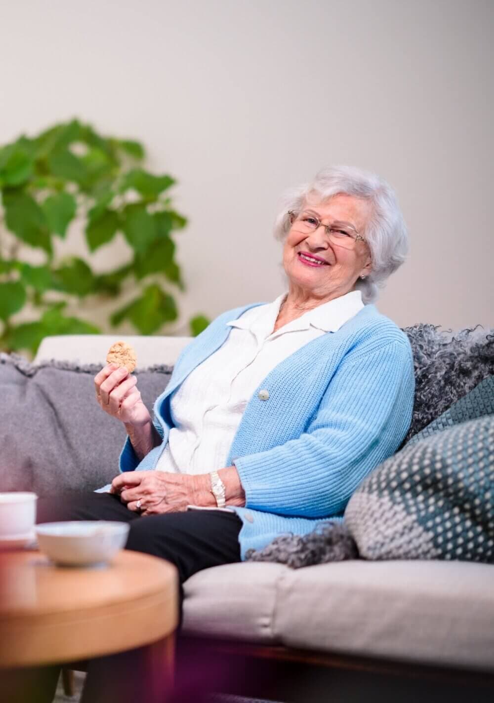 Elderly woman with white hair and glasses, wearing a blue sweater, smiling while holding a cookie, seated on a couch. - Home Instead