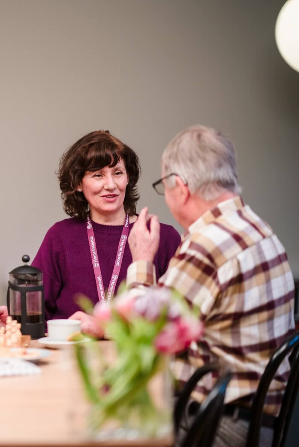 Two people having a conversation at a table with a French press and coffee cups, in a casual indoor setting. - Home Instead