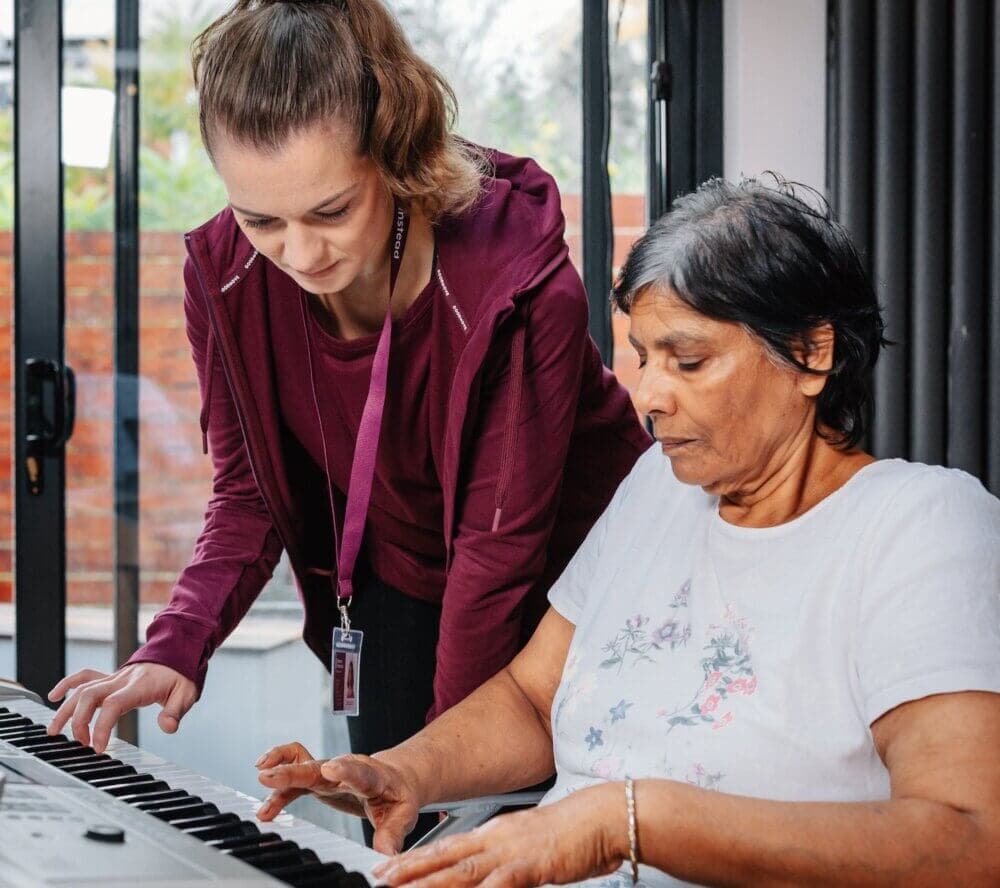 A woman assists another woman in playing the keyboard in a sunny room with large windows. - Home Instead