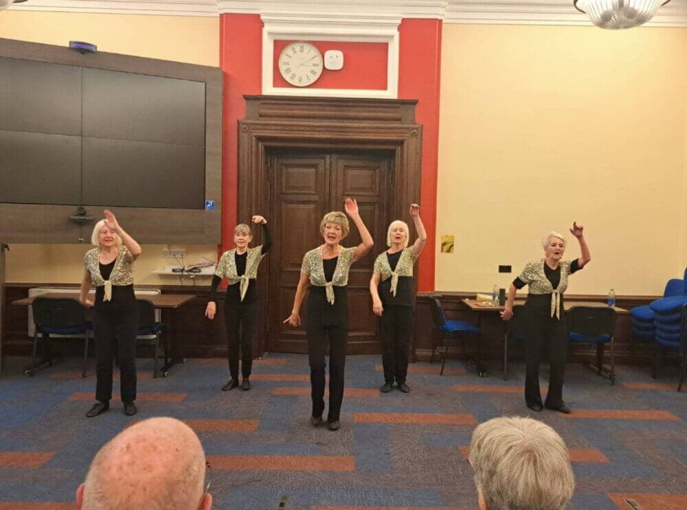 Five women performing a dance routine in a room with chairs, a large screen, and a clock on the wall. Audience heads visible. - Home Instead