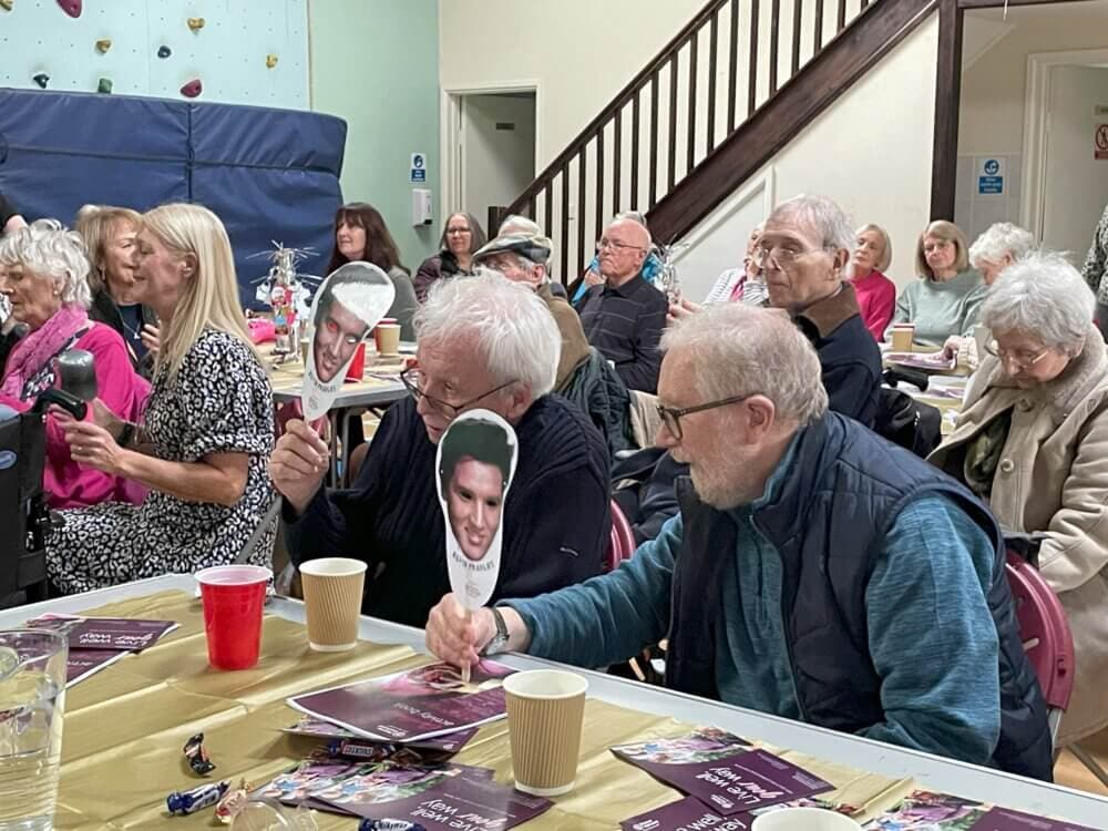 A group of elderly people seated at tables holding masks with a young man's face at a community event. - Home Instead