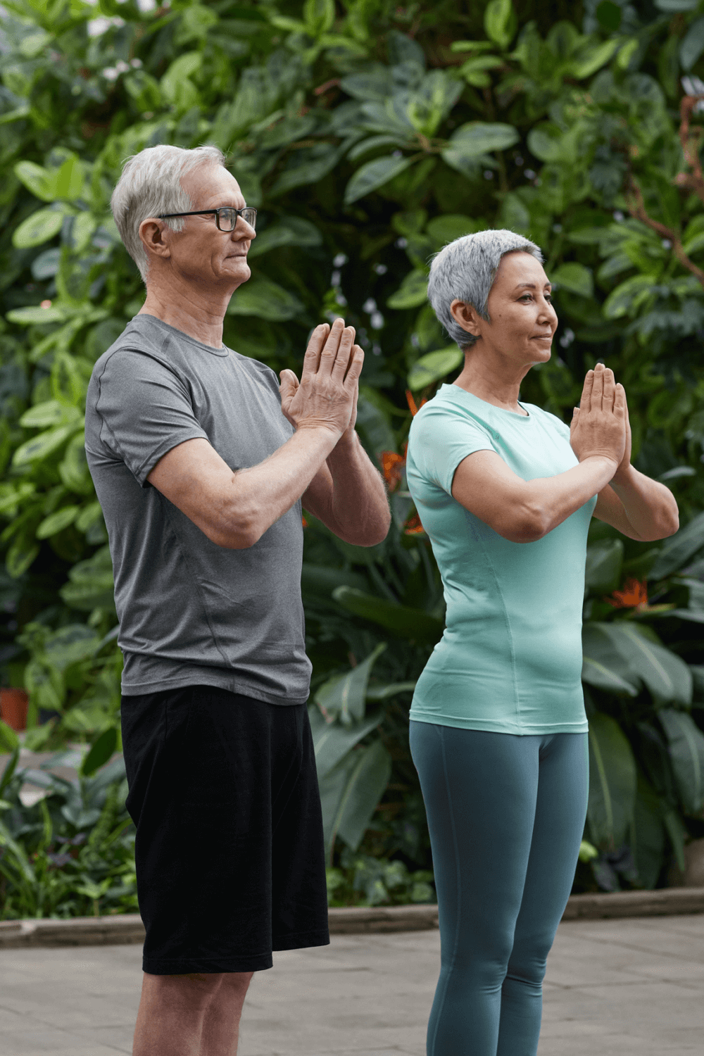 Two older adults practice yoga outdoors, standing in a namaste pose with hands together, surrounded by lush green plants. - Home Instead