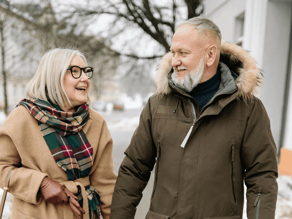 An older couple, both wearing winter coats, smile and hold hands while walking outside on a snowy day. - Home Instead