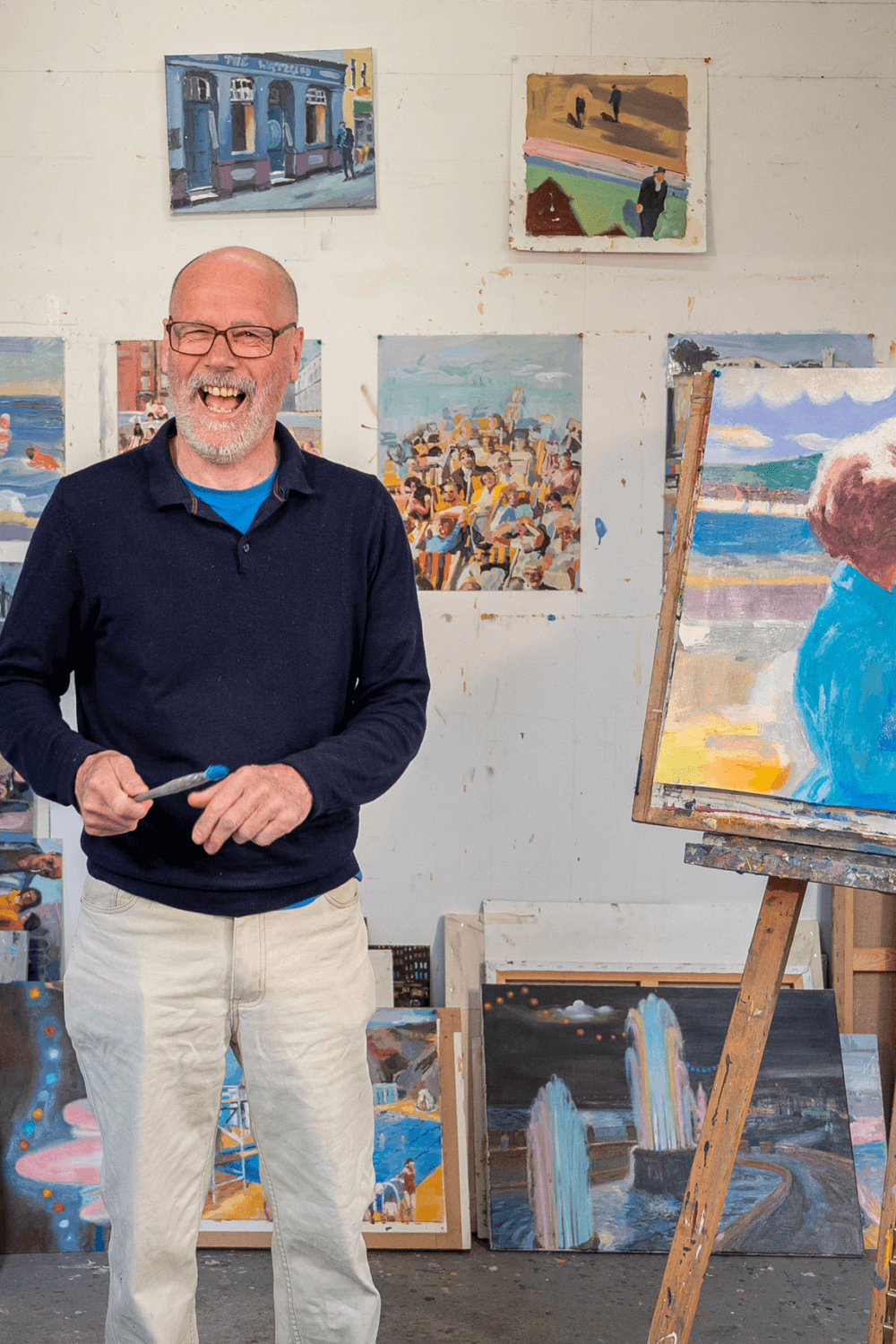 A man stands smiling in an art studio, surrounded by colorful paintings, with one displayed on an easel. - Home Instead