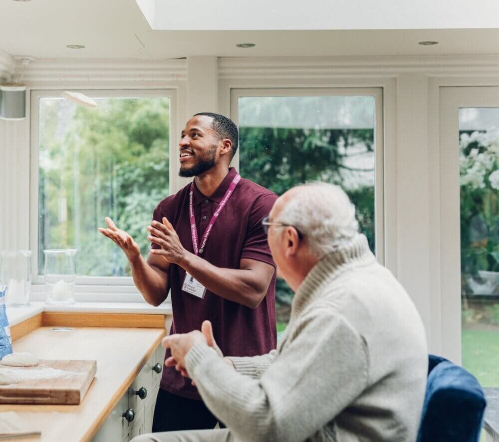 A caregiver and an elderly man converse happily in a bright kitchen with large windows and a lush view outside. - Home Instead