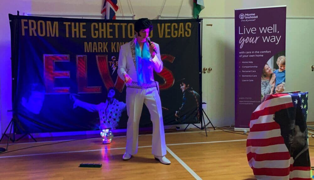 A performer in a white jumpsuit sings on stage with "From the Ghetto to Vegas" and "Elvis" banners in the background. - Home Instead