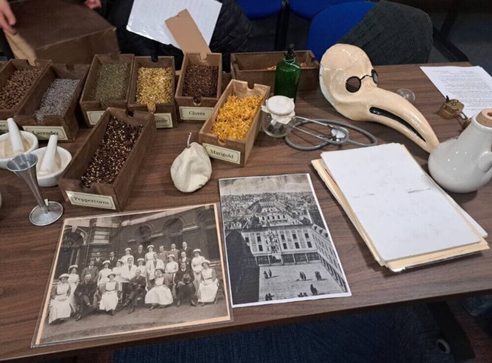 A table with herbs, medical tools, historical photos, and a plague doctor's mask arranged on top. - Home Instead