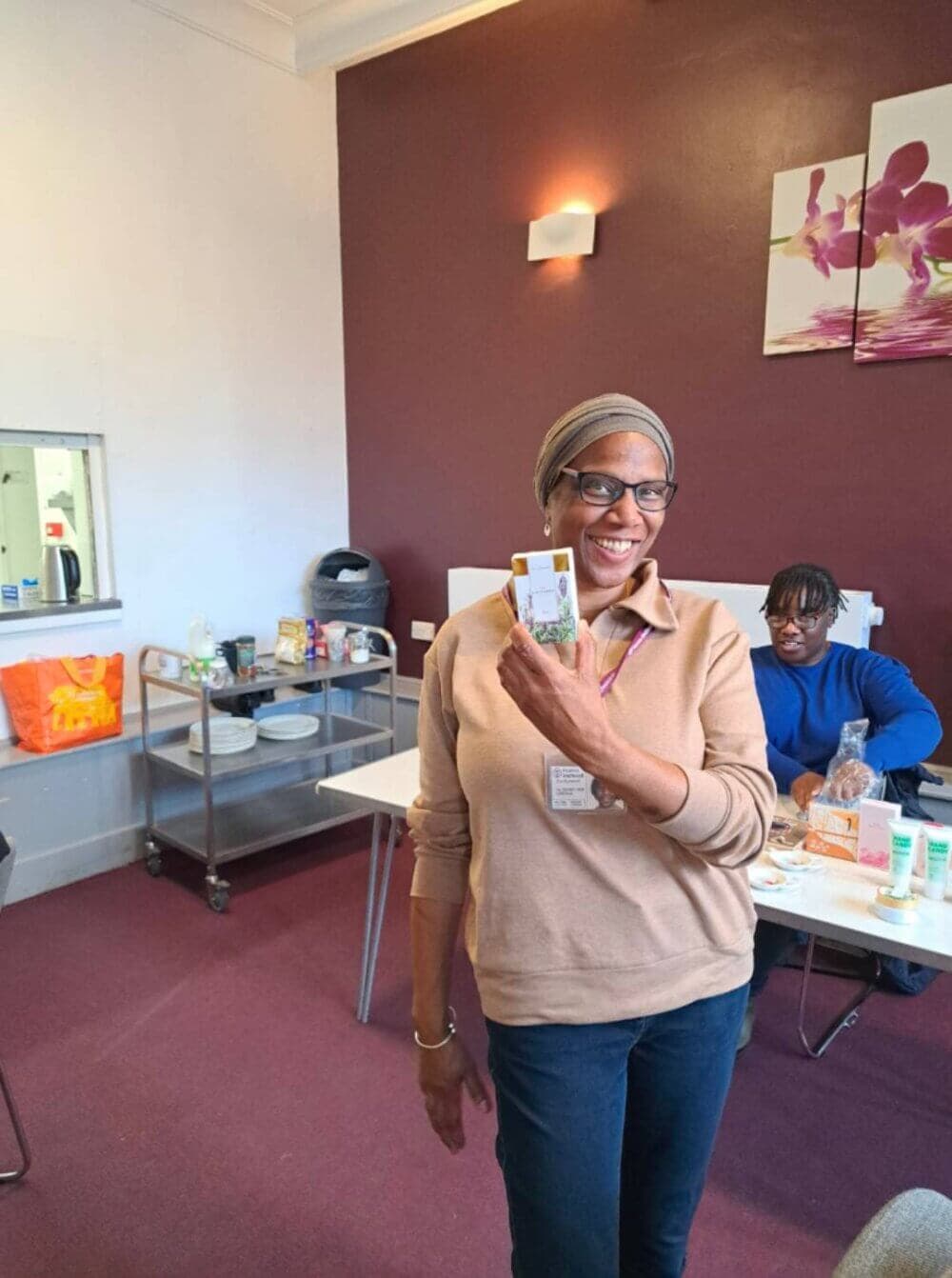 A smiling woman in a beige sweater holds up a small item, standing in a room with a red wall and various supplies. - Home Instead