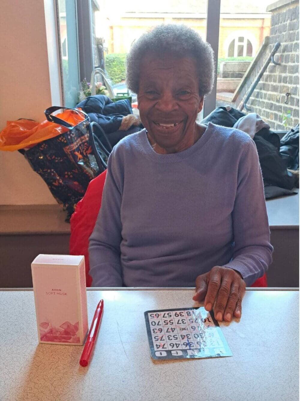 Elderly woman sitting at a table with a bingo card and pen, smiling warmly at the camera, with a window in the background. - Home Instead
