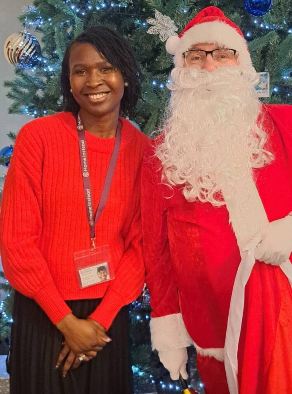 A smiling person in a red sweater stands beside Santa Claus in front of a decorated Christmas tree. - Home Instead