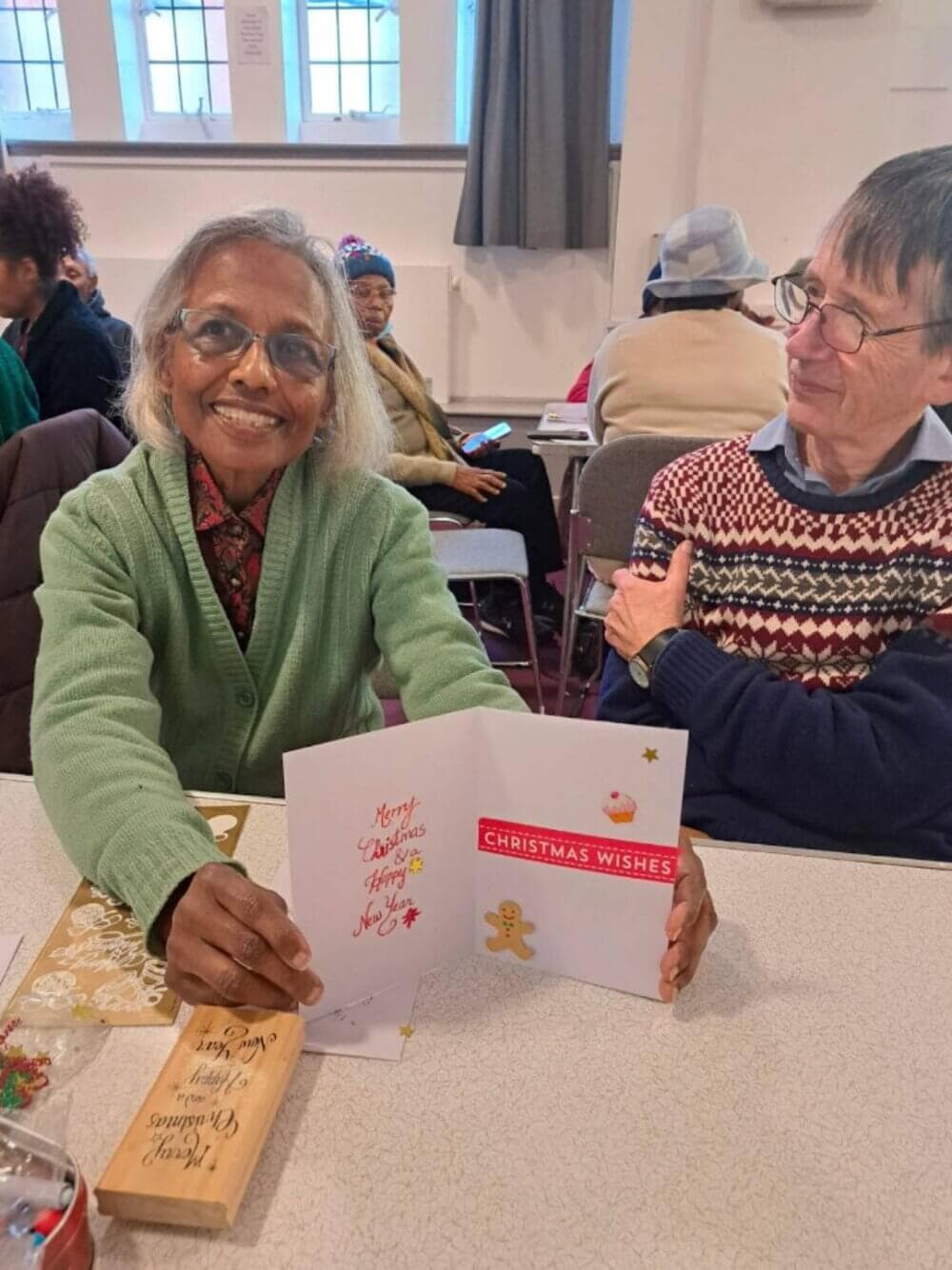 Two elderly individuals sitting at a table in a community setting, holding up a Christmas card and smiling. - Home Instead