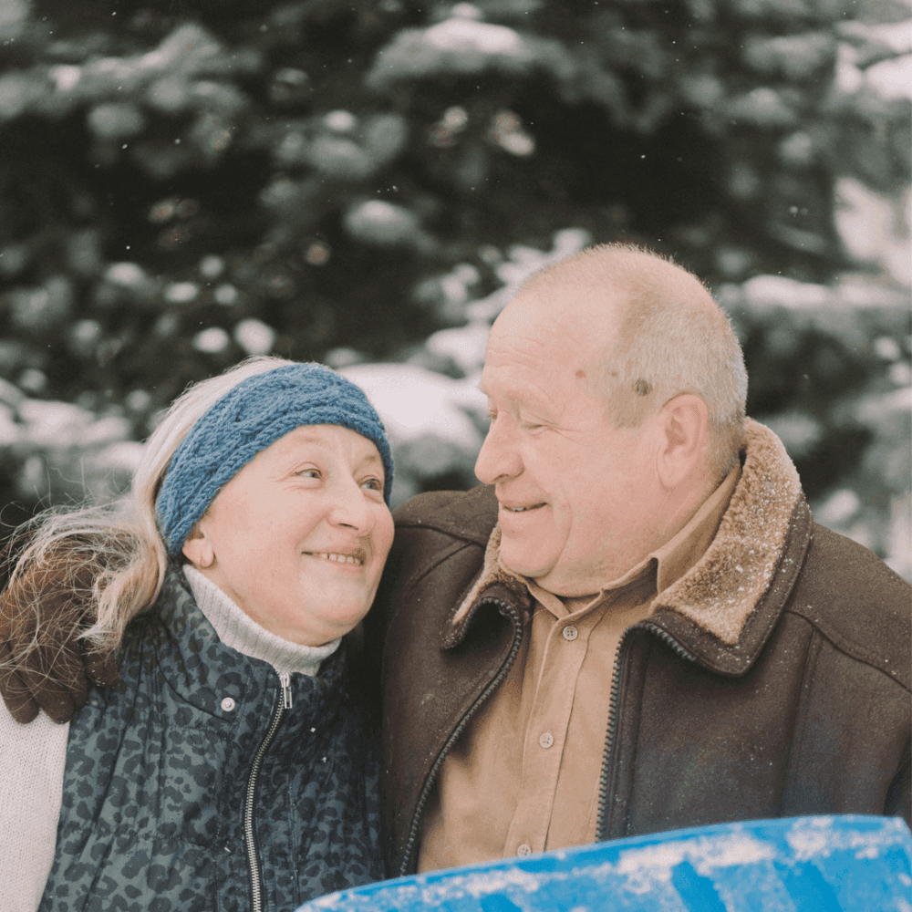 An elderly couple warmly dressed, smiling and embracing each other in a snowy outdoor setting. - Home Instead