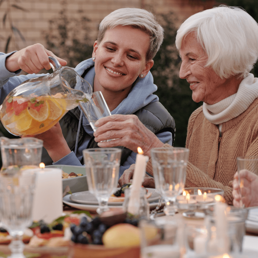 Two women enjoying an outdoor meal. One pours a drink while the other holds a glass, with food and candles on the table. - Home Instead