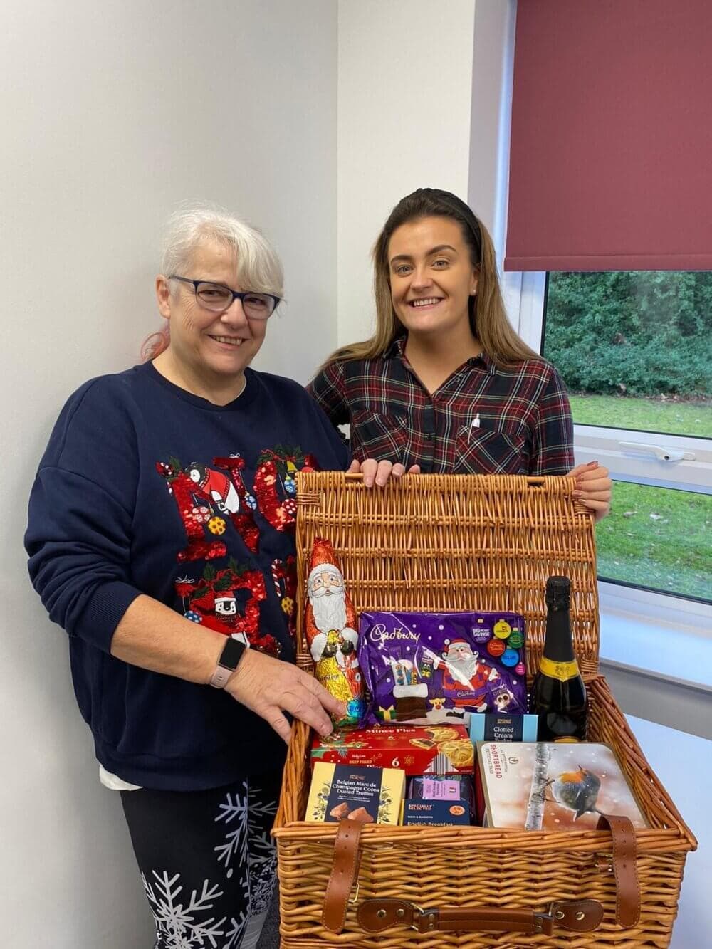 Two women smile while standing, holding a wicker basket filled with festive treats and gifts placed on a table. - Home Instead
