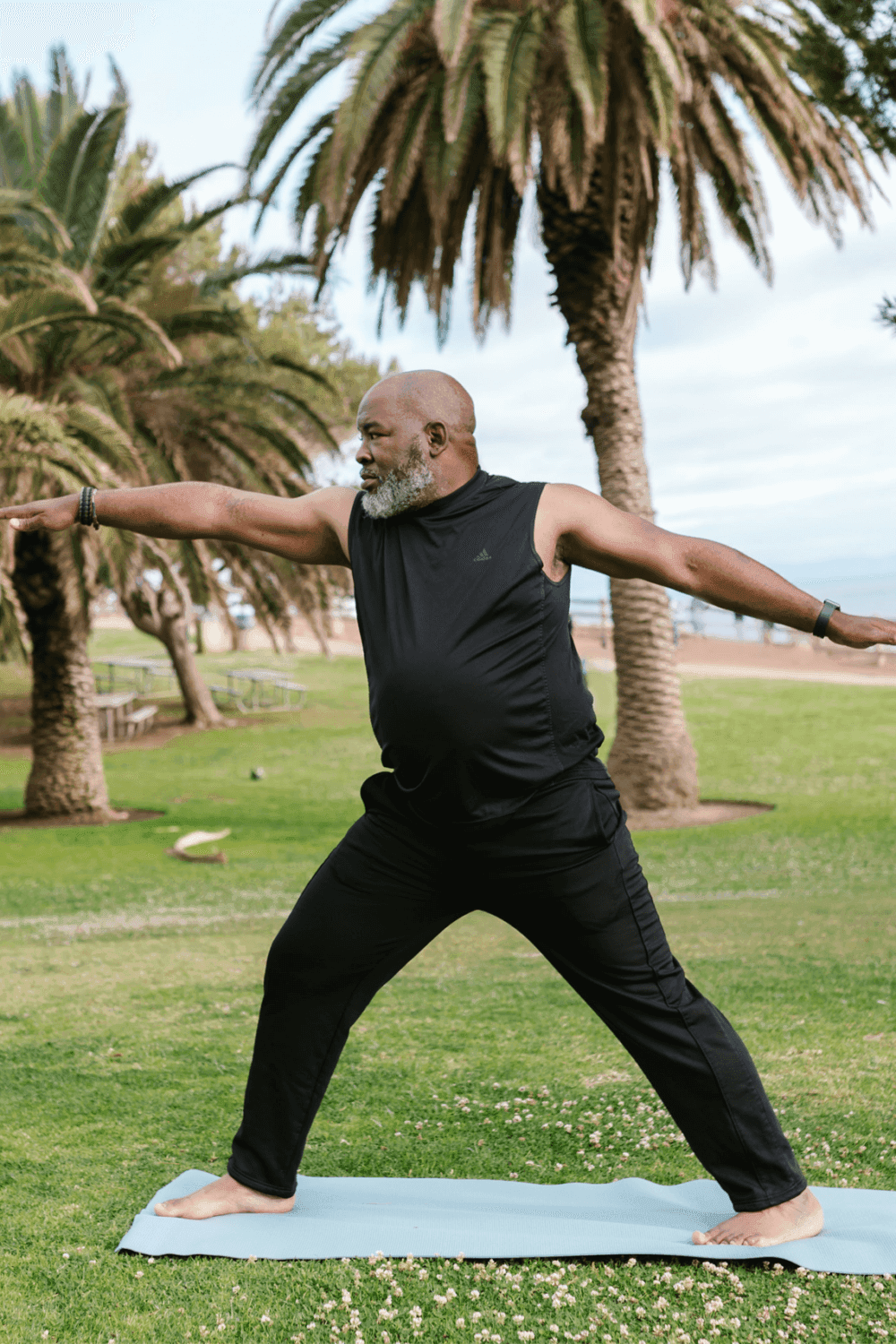 A man practices yoga in a park, performing a Warrior pose on a blue mat with palm trees in the background. - Home Instead