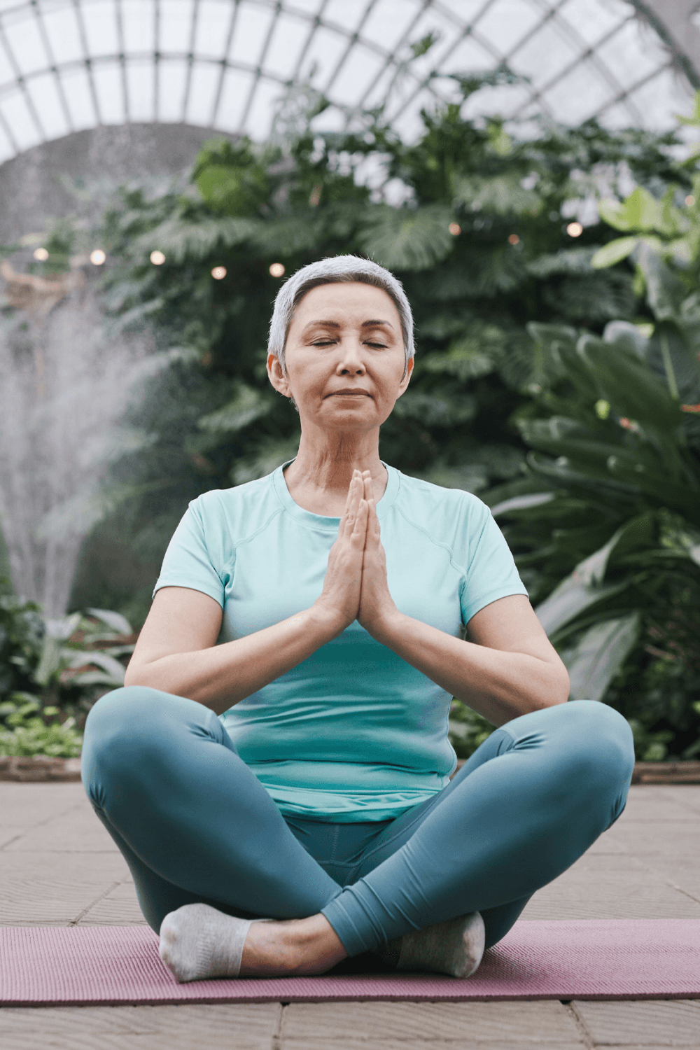 A woman wearing a light blue outfit is meditating in a lotus position on a yoga mat in a greenhouse. - Home Instead