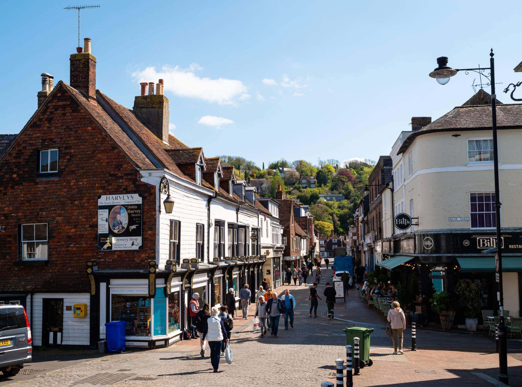 Busy street in a charming town, with old brick buildings, people walking, and a hilly background with greenery. - Home Instead