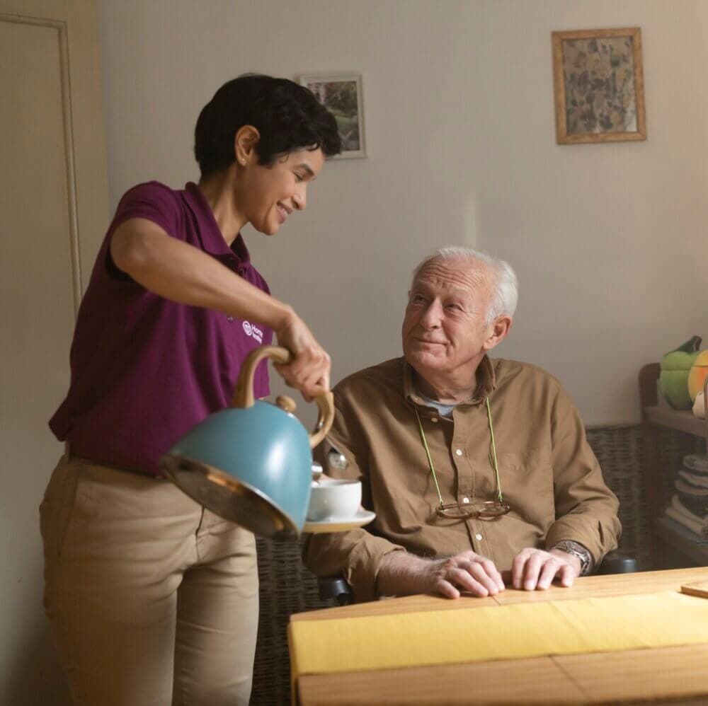 A caregiver in a purple shirt pours tea for an elderly man sitting at a table, smiling and engaging with him. - Home Instead