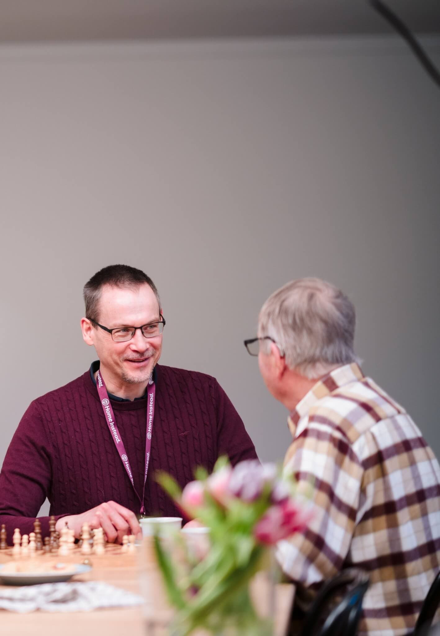 Two men, one in a maroon sweater, the other in a checkered shirt, chatting and playing chess at a wooden table. - Home Instead