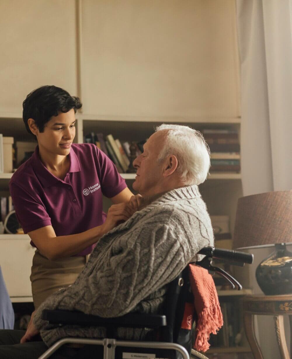 A caregiver helps an elderly man, who is sitting in a wheelchair, adjust his sweater in a cozy, book-filled room. - Home Instead