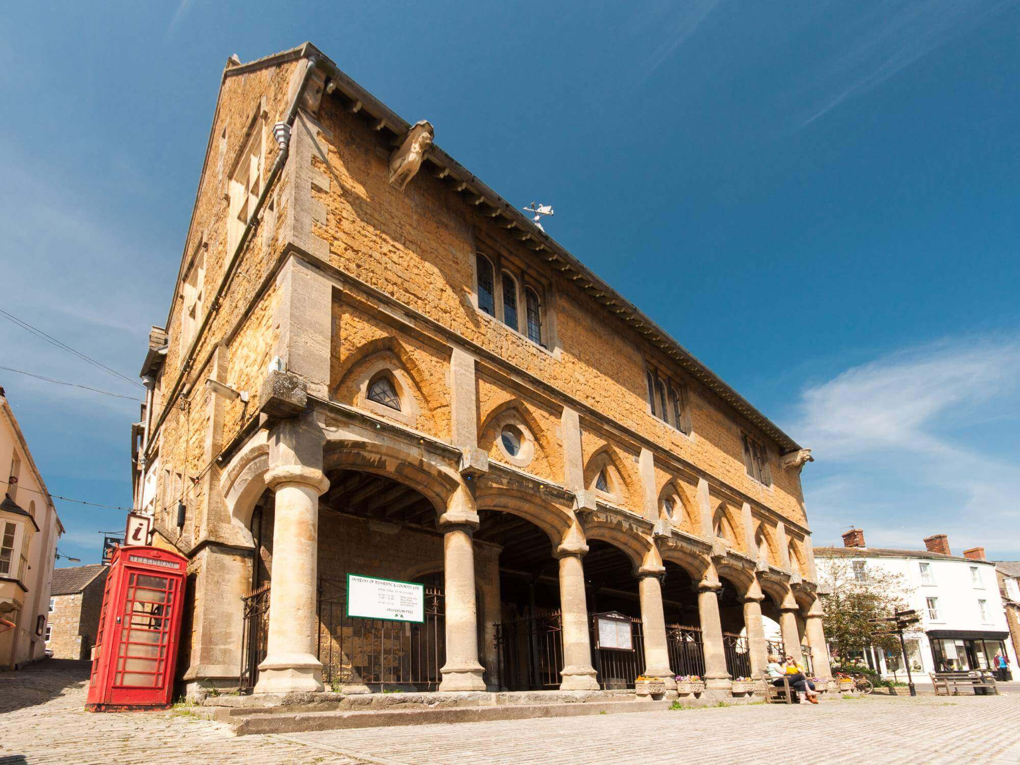 Historic stone market hall with arched entrances and columns under a clear blue sky, red phone booth to the left. - Home Instead