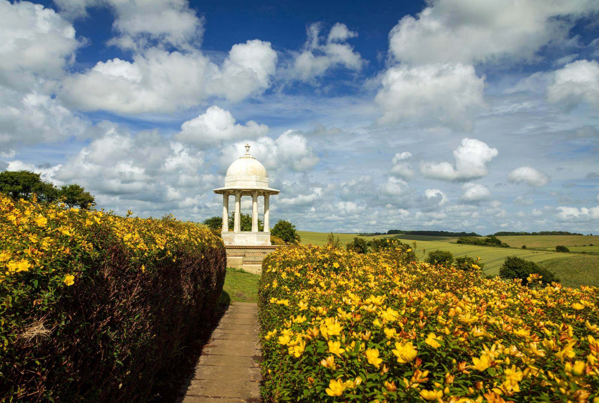 A small white domed pavilion amidst a scenic landscape with yellow flowers, green fields, and a partly cloudy sky. - Home Instead