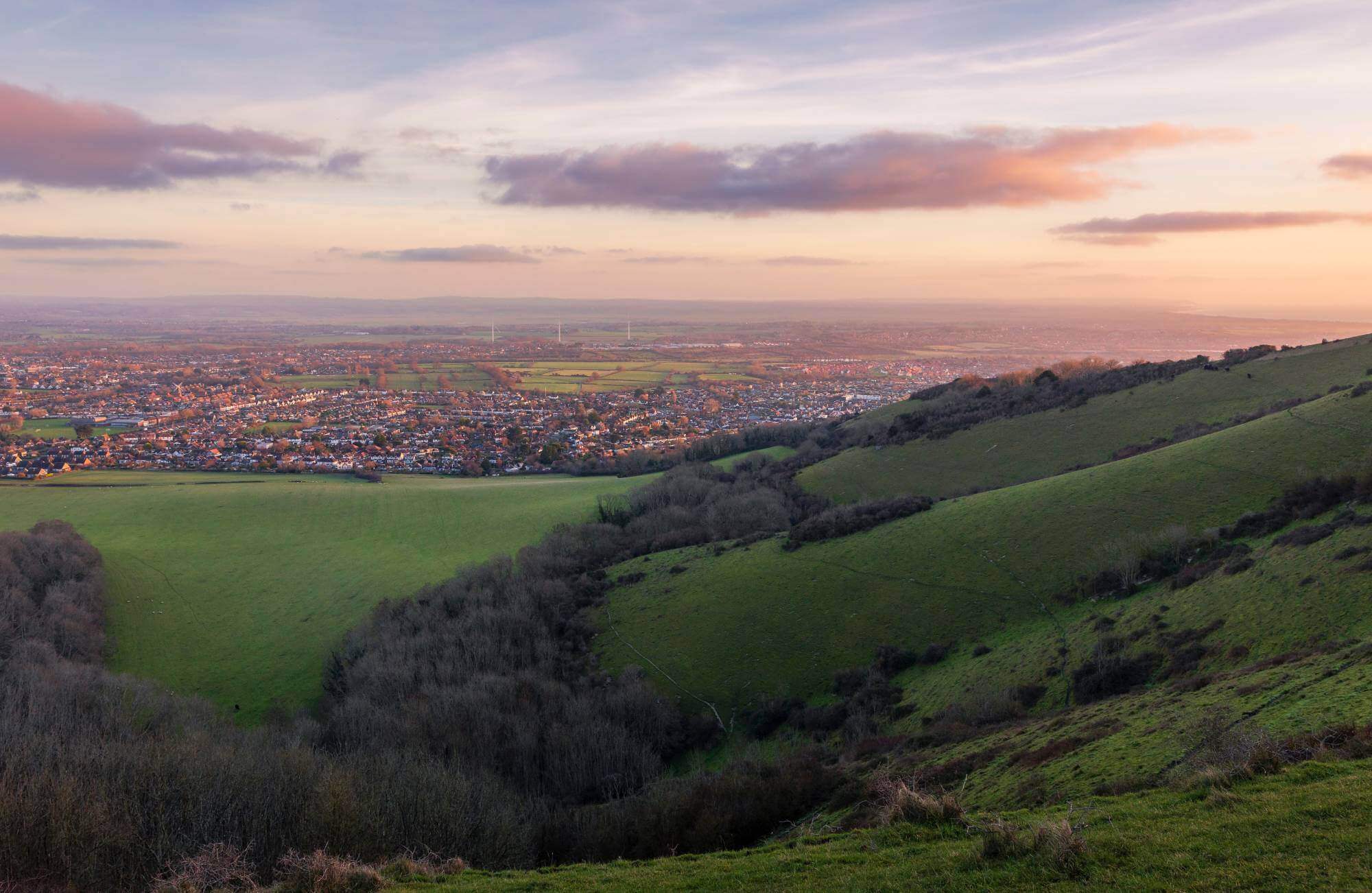 Panoramic view of a cityscape at dusk, seen from a green hillside with a cloudy sky and rolling hills in the foreground. - Home Instead