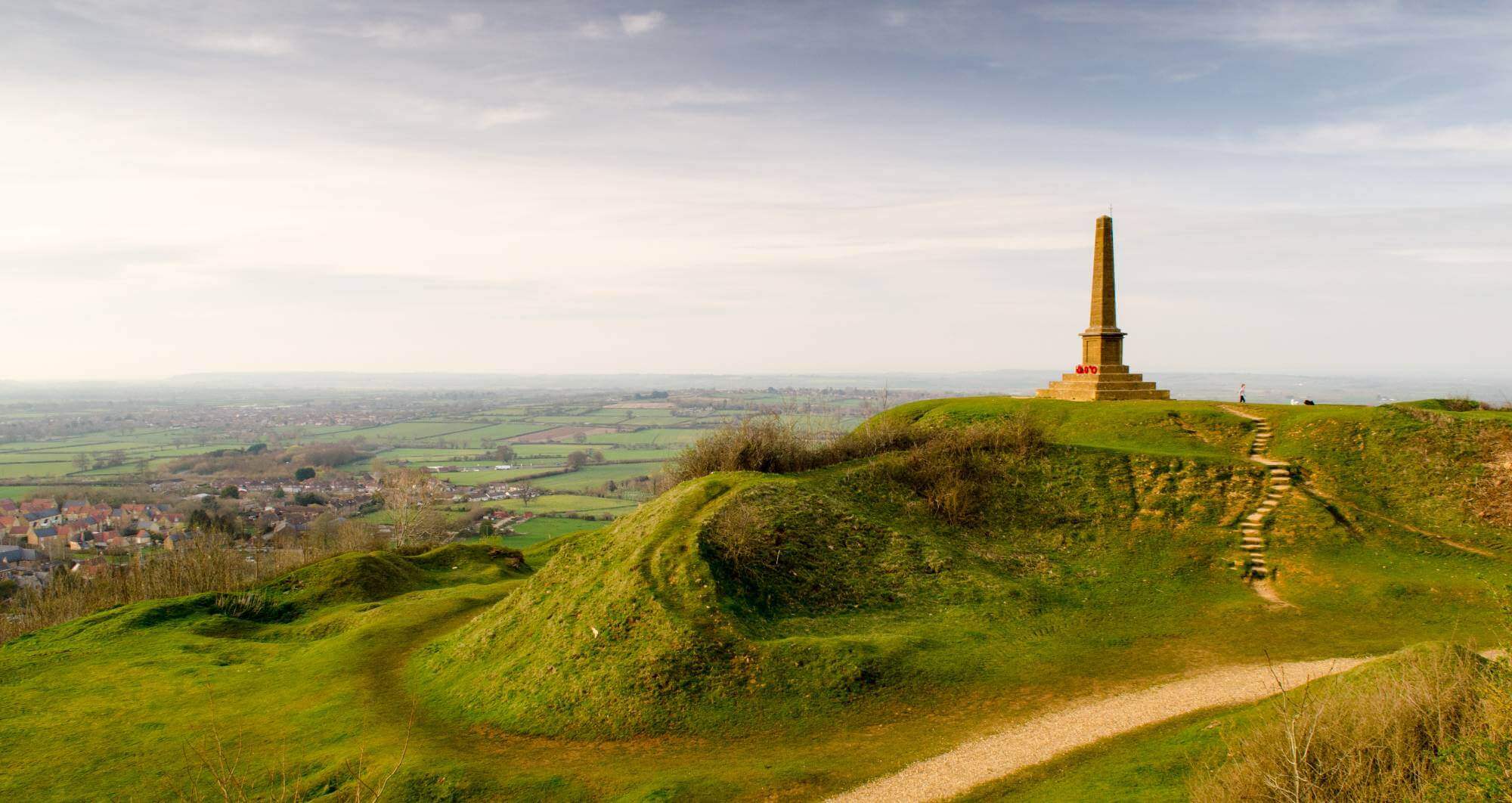 A tall monument on a grassy hilltop overlooking a vast landscape with distant fields and a few scattered buildings. - Home Instead