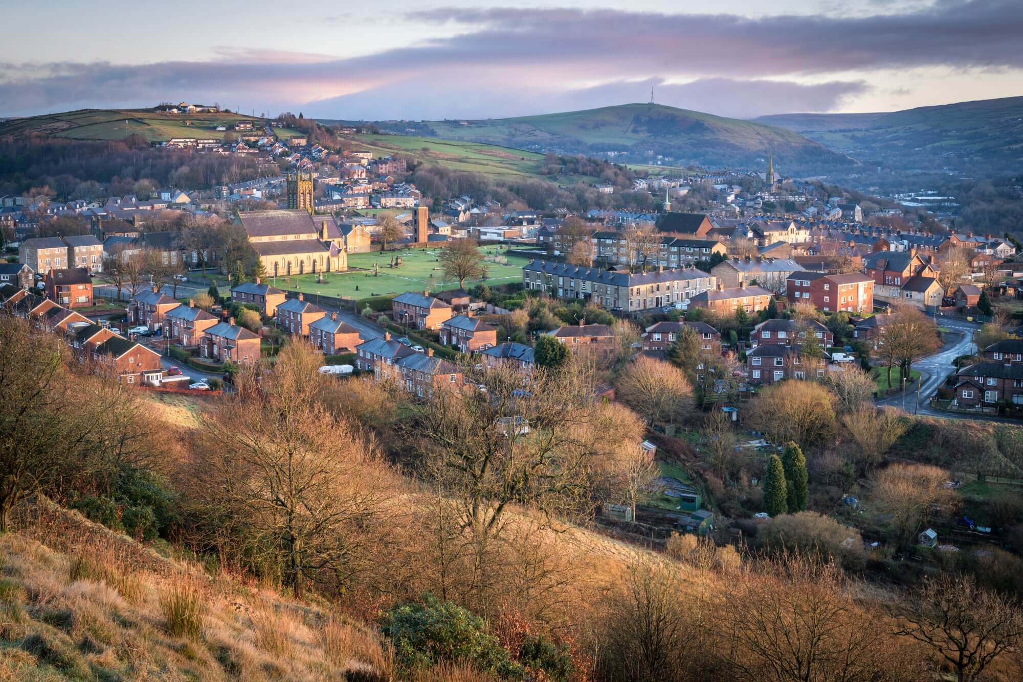 A scenic view of a small town with houses, a church, and rolling green hills under a partly cloudy sky. - Home Instead