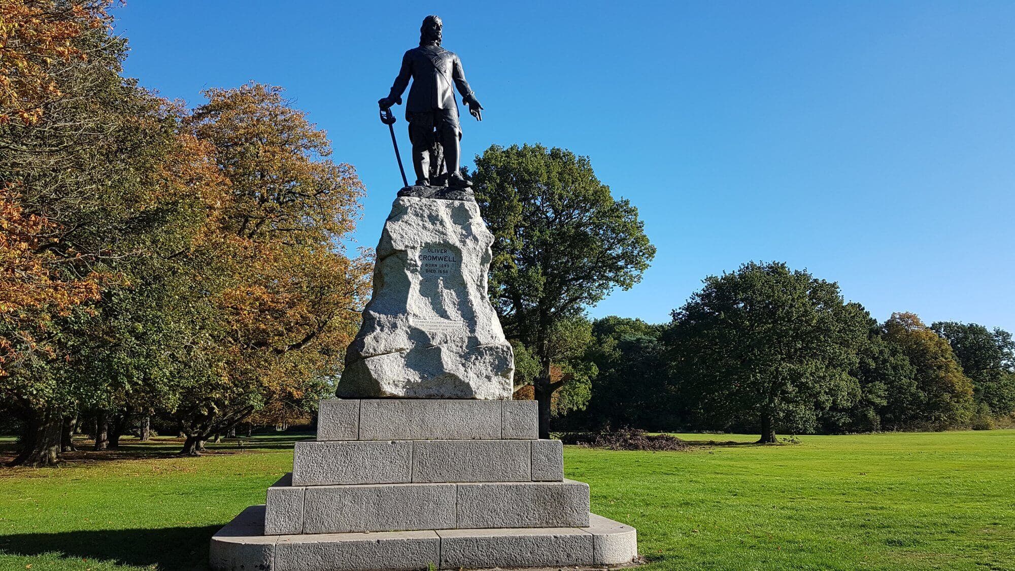 Statue of a standing figure on a stone pedestal surrounded by trees in a park under a clear blue sky. - Home Instead